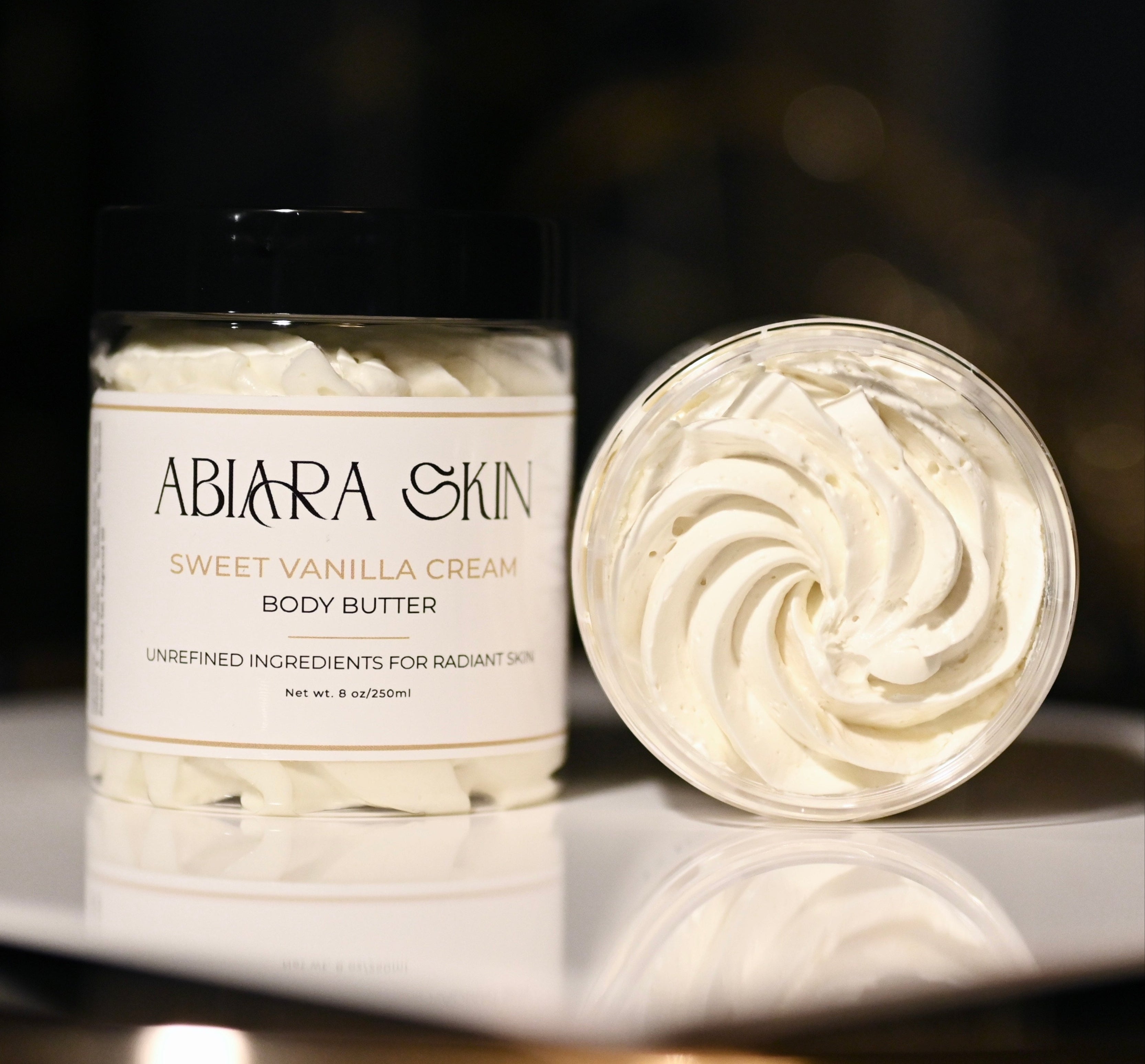 ABIARA SKIN (formerly Body Essentials by Abiara) Sweet Vanilla Cream body butter jar and container on a reflective surface with a dark background