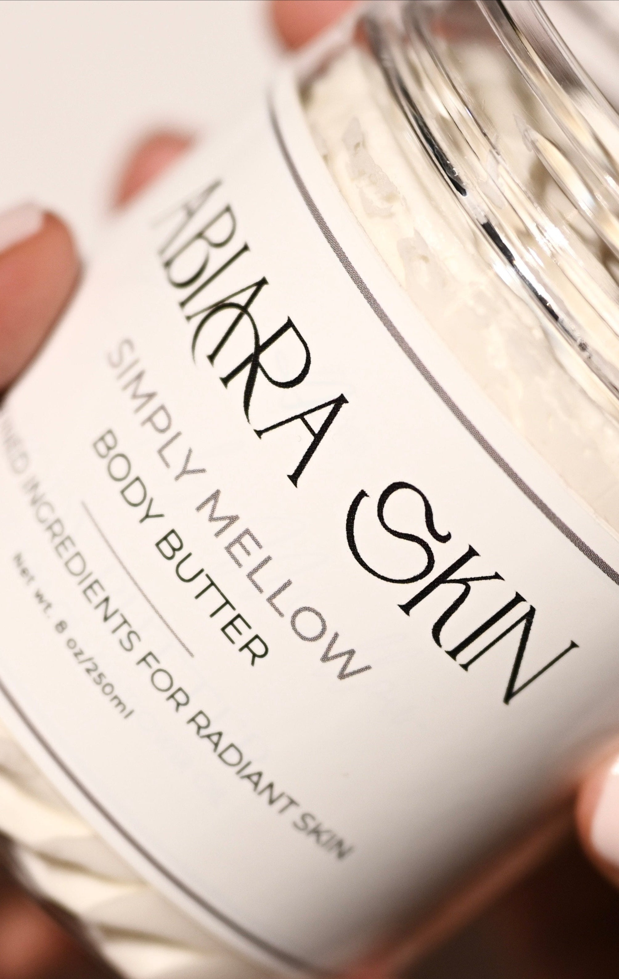Abiara Skin Simply Mellow Body Butter jar held by a hand with light pink nail polish.