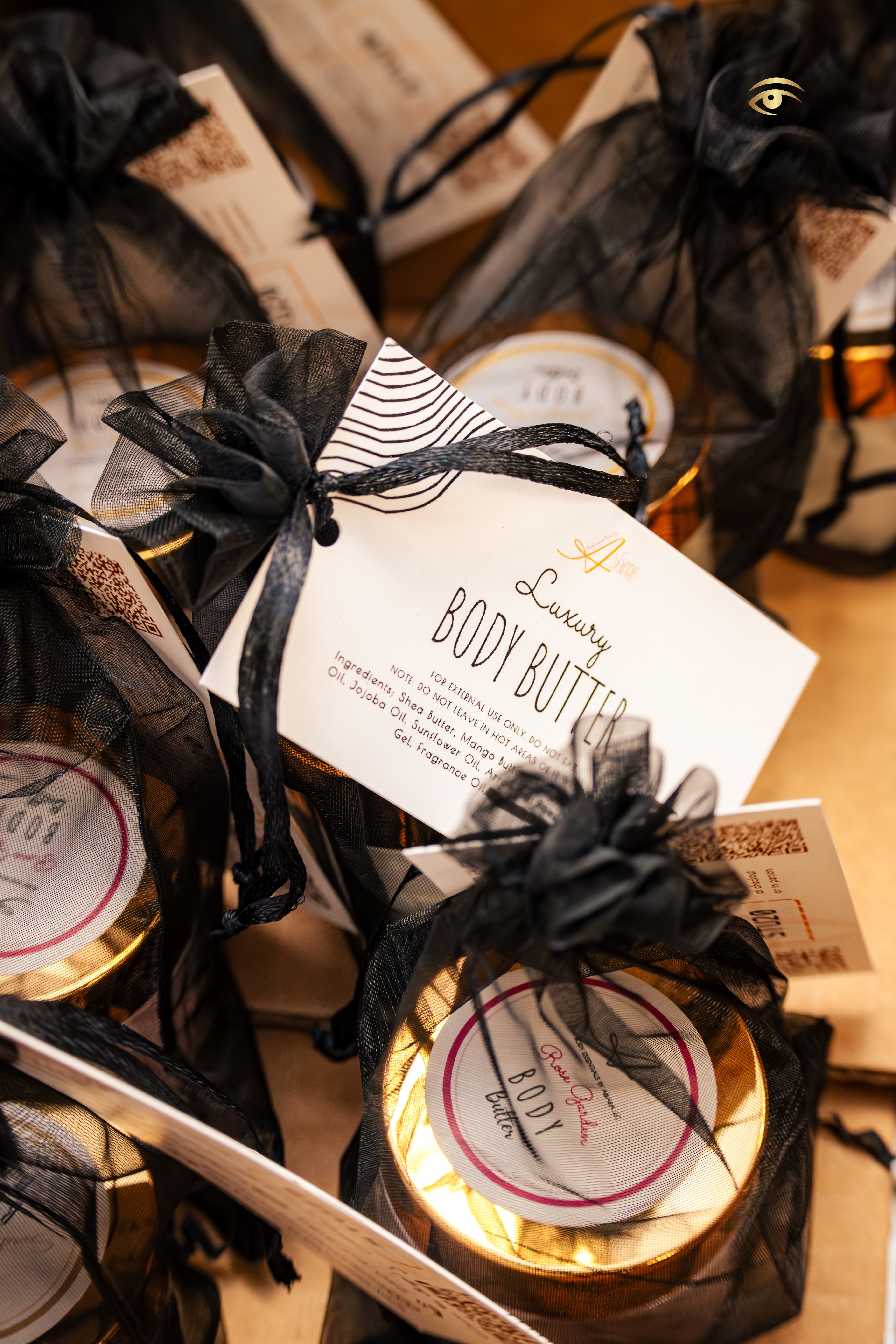 Close-up of Abiara™ Skin luxury body butter jars packaged in black organza gift bags with gold lids and custom labels, accompanied by branded ingredient cards, arranged for party favor presentation.